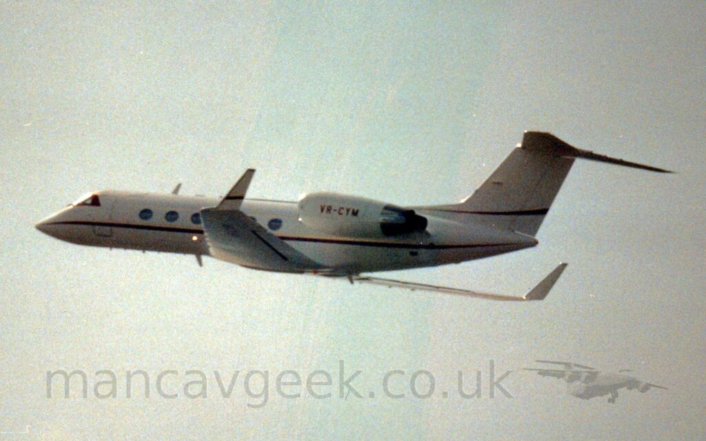Side view of a twin engined BizJet flying from left to right at a low altitude, with undercarriage and flaps retracted, and the nose raised sharply, suggesting it has just taken off. The plane is white, with a thin red stripe running along the body, under the oval passenger cabin windows, as well as along the bottom of the tails, and the sides of the up-turned wingtips. The red registration "VR-CYM" is on the sides of the white engine pods mounted on the sides of the rear fuselage. White clouds fill the background, with patches of blue threatening to peek through in the middle.