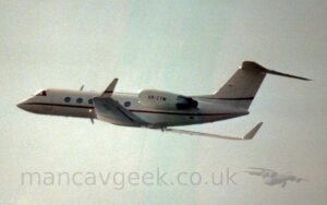 Side view of a twin engined BizJet flying from left to right at a low altitude, with undercarriage and flaps retracted, and the nose raised sharply, suggesting it has just taken off. The plane is white, with a thin red stripe running along the body, under the oval passenger cabin windows, as well as along the bottom of the tails, and the sides of the up-turned wingtips. The red registration "VR-CYM" is on the sides of the white engine pods mounted on the sides of the rear fuselage. White clouds fill the background, with patches of blue threatening to peek through in the middle.