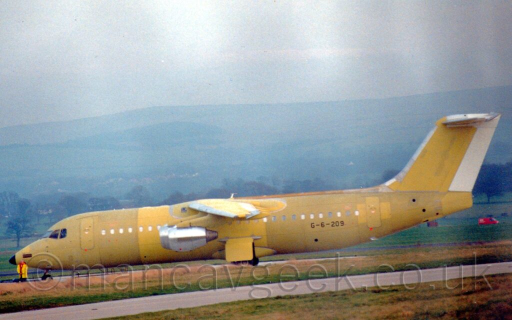 Manchester, Woodford, EGCD, BritishAerospace, BAE146, OnThisDay, AvGeek, aviation, planespotting, photography Side view of a high-winged, 4 engined jet airliner parked facing to the left. The plane is mostly yellow, with various barrel sections of the fuselage and wings and different panels painted in different shades. The engine pods are bare metal, and the rudder on the tail is plain white. The black registration "G-6-209" is on the lower rear fuselage. Green grass lines a taxiway in the foreground, with more grass leading up to trees marking the edge of the airfield in the background beyond that, and rolling hills in the distance, under grey skies.