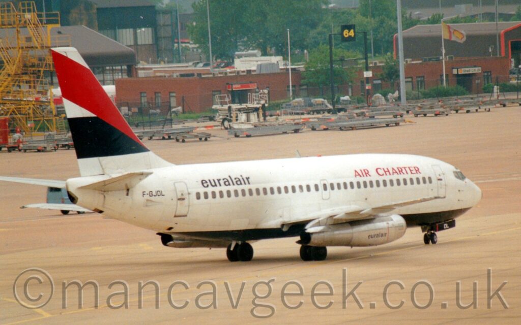 Side view of a white, twin engined jet airliner taxiing from left to right. The plane is mostly white, with a black belly, and red "Air Charter" titles on the upper forward fuselage, and black "EuralAir" titles on the upper rear. There is a thick black stripe across the tail, overlaid with a thick diagonal red band. The black registration "F-GJDL" is on the rear fuselage, under the tail. Grey concrete apron fills most of the foreground and part of the background, with rows of grey freight trolleys lined up in the distance, with brown brick buildings and trees beyond that.