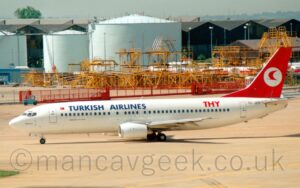 Side view of a twin engined jet airliner taxiing from right to left. The plane is mostly white, with large blue "TurkishAirlines" titles on the upper forward fuselage, red letters "THY" on the upper rear, and the black registration "TC-JDY" under the tail.. The tail is red, with a stylised red flying bird in a white circle. Grey concrete apron fills the foreground, with a collection of bizarre-looking yellow metal frames in the background, in front of so duck-egg blue fuel tanks. Black hangars stretch across the frame beyond that, under typically grey Mancunian sky.