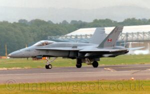 Side view of a twin engined, single seat, military fighter jet taxiing from right to left along a black runway. The plane is dark grey on top and a lighter grey on the bottom, with the very dark grey number "748" on the nose just in front of the cockpit canopy, and the light grey word "Canada" on the upper fuselage just behind the cockpit. The outward-canted twin tails are dark grey on the inner surfaces, and at the front of the outer surfaces, with the rest being the same light grey as the lower fuselage. A small red and white Canadian flag is in the middle of the outer face of the tail, above the dark grey number "188748". Below the cockpit canopy there is the dark grey outline of a circle, containing a dark grey image of a maple leaf, the symbol of Canada, as well as curved black shape meant to replicate the shape of the cockpit canopy, making it harder for someone seeing it fly to tell which way up it is. Grass at the bottom of the frame lines the side of the runway, with more grass on the far side leading up to a large, white and grey hangar on the right of the frame, and trees on the left, under flat grey sky.