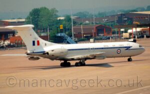 Side view of a white, 4 engined jet airliner with the engines mounted in pairs on the sides of the rear fuselage, taxiing from left to right. The plane has white top surfaces, and grey lower surfaces, with a blue stripe running along the body, covering the passenger cabin windows. The engine pods are grey, with the serial "XV102" in black. There is a red, white, and blue roundel on the lower forward fuselage, and a blue, white, and red fin flash on the tail, under the number "102". There are black "Royal Air Force" titles on the upper forward fuselage. The plane is carrying a pair of air-to-air refuelling pods, one under each wing, allowing it to dispense fuel to allied aircraft. Grey concrete apron fills the foreground, with a collection of brown buildings nestled in trees in the background, under dreary grey skies.
