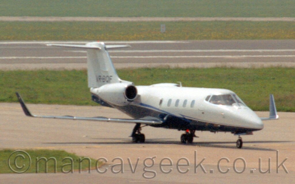 Forward side view of a twin engined BizJet taxiing from left to right along a grey taxiway. The plane is white on top and towards the front, and dark blue underneath and to the rear. White engine pods are mounted on the upper rear fuselage. The black registration "VR-BQF" is on the bottom of the white tail. The large, up-turned wingtips are also dark blue. A pair of grey taxiways fill the foreground, with a strip of green grass on the left separating them until the point where they join. More grass in the background lead up to a black runway, with more grass filling the top of the frame on the far side.