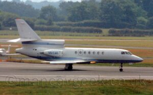 Side view of a 3 engined BizJet moving from left to right. The plane is mostly white, with 3 thin blue stripes running along the lower body, and another 3 stripes running along the bottom of the centre engine air intake and tail. The blue registration "VR-CAE" is on the side of the engine pods mounted on the sides of the rear fuselage. Green grass fills the foreground, with more grass and taxiways in the background leading up to trees in the distance.