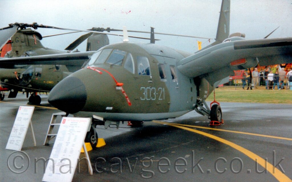 Three-quarter front view of a gull-winged, twin propellor-engined military transport aircraft parked on a black asphalt taxiway, on static display at an airshow. The plane has a dark grey and olive-green camouflage on the top and sides of the body and wings, with a light grey belly and undersides of the wings. The grey numbers "303-21" are on the side of the fuselage, under the windows at the sides of the cockpit, with a black nose-cone. White boards giving text information about this plane are propped up in front of the plane, the writing on them visible but incomprehensible. A strip of green grass visible under the wing on the right of the frame leads up to crowds of people in the background, with a twin-rotor helicopter in the background on the left, under flat grey sky.