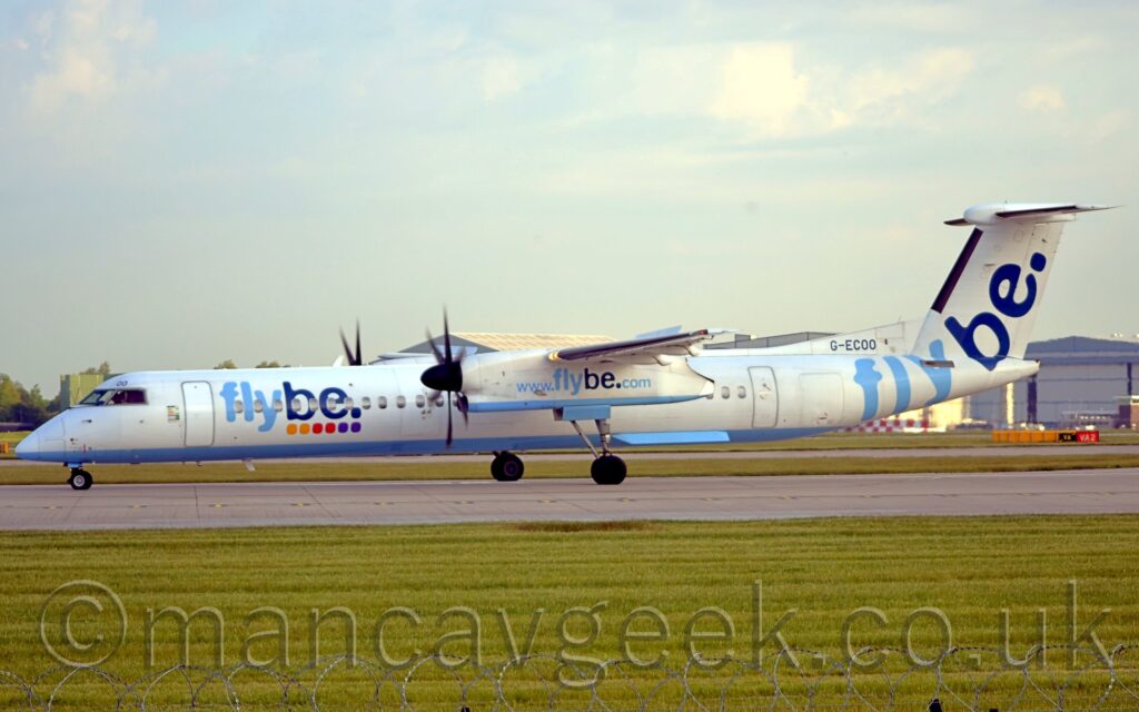 Side view of a high-winged, twin propellor-engined airliner parked on a grey runway. The plane is mostly white with a light blue belly, with light and dark blue "FlyBe" titles over a series of coloured dots (2 yellow, 2 red, 2 purple) on the side of the forward fuselage, and the black registration "G-ECOO" on a fillet at the front of the tail. A larger, diagonal version of the "FlyBe" logo appears on the rear fuselage and tail, and the light blue text "www.flybe.com" on the side of the engine pods under the wings. Green grass fills the foreground, and in the background leads off to a large grey hangar on the right of the frame, and trees on the left, under flat white sky.