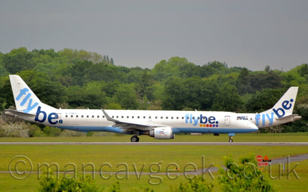 Side view of a twin engined jet airliner moving from left to right along a runway. The plane is largely white, with a light blue belly, and light and dark blue "FlyBe" titles on the sides of the forward fuselage, over 6 coloured dots - 2 yellow, 2, red, and 2 purple. This logo is repeated on the sides of the white engine pods under the wings, with additional light blue "[dot]com" at the end. The black registration "G-FBEF" is on the upper rear fuselage, with light and dark blue "FlyBe" titles on the tail and rear fuselage. Green grass fills the foreground, with bushes poking into frame right at the bottom. In the background, a near identical plane with the registration "G-FBJH" on the upper rear fuselage is taxiing to the left, partially hidden behind this plane, with trees beyond that, under grey sky.