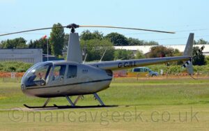 Side view of a silvery-grey helicopter parked facing to the left on a grey concrete pad surrounded by green grass. There is a black stripe running along the body, with small text "R44 Raven II" behind the rear cabin doors, and the black registration "G-CJLL" on the silver tail boom. The horizontal rotor blade on the tale has black and white bands to make it more visible, especially when spinning. Green grass fills the foreground, with trees and grey buildings in the background, under blue-grey skies.