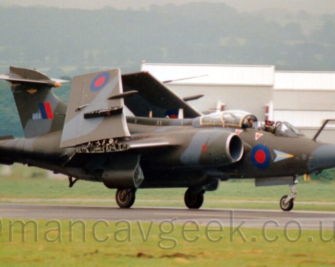 Side view of a twin jet-engined, military light bomber taxiing from left to right along a black runway with it's wings folded up. The plane is covered in an olive-green and grey camouflage, with blue and red roundels on the lower forward fuselage, under the cockpit, and under the outer wing panels, the latter visible because the wings are raised into their stowed position. There is a blue and red fin flash on the tail, next to the grey number "864". The cockpit canopy is in the slid back position, with both crew members visible in their tandem seats. Green grass lines the runway in the foreground, with a large grey building dominating the background on the right, with rolling, tree covered hills fading into the hazy grey sky.