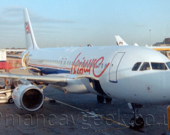 Front side view of a twin engined jet airliner parked facing to the right, nose-in to it's gate, as seen from inside the terminal building. The plane is largely white, with large, red, apparently hand written "Leisure" titles on the forward fuselage and a thick blue strip running along the body under a thin red pinstripe. The black registration "G-UKLJ" is on the upper rear fuselage, with the last two letters, LJ, repeated on the open nose-gear doors. The tail is white, with half of a waving UK flag in the middle. The bed of a white and pink catering truck is raised to the open rear door of the plane, with a low, white, baggage belt by an open cargo door in the belly just behind the wings, and another cargo door open in front of the wings. In the background, on the right, a brown and cream terminal building stretches across the frame from the right, with a couple of planes parked in front, with pale blue sky filling the rest of the frame.