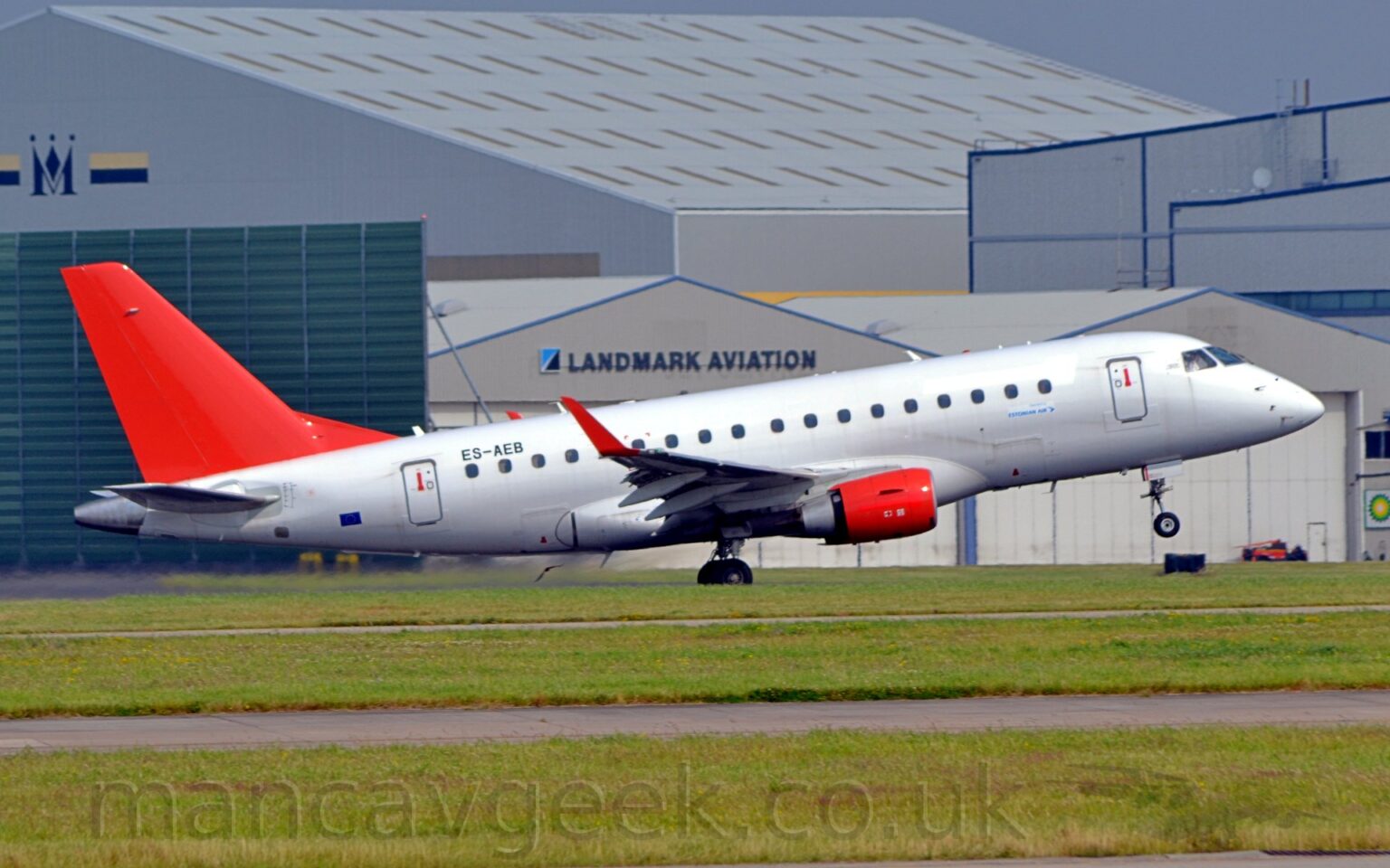 Side view of a twin engined jet airliner moving from left to right at high speed, with flaps extended from the rear of the wing and the nose-wheel raised sharply, suggesting it is in the middle of taking off. The plane is almost entirely white, with plain red tail and engine pods, mounted under the wings. There are small, blue, "Estonian Air" stickers un the first 2 passenger cabin windows, while the black registration "ES-AEB" is over the last 2. The up-turned wingtips are also bright red. Green grass lines taxiways in the foreground, while a selection of grey hangars line the background, under grey sky.