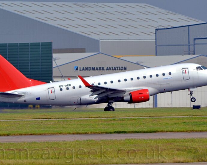 Side view of a twin engined jet airliner moving from left to right at high speed, with flaps extended from the rear of the wing and the nose-wheel raised sharply, suggesting it is in the middle of taking off. The plane is almost entirely white, with plain red tail and engine pods, mounted under the wings. There are small, blue, "Estonian Air" stickers un the first 2 passenger cabin windows, while the black registration "ES-AEB" is over the last 2. The up-turned wingtips are also bright red. Green grass lines taxiways in the foreground, while a selection of grey hangars line the background, under grey sky.