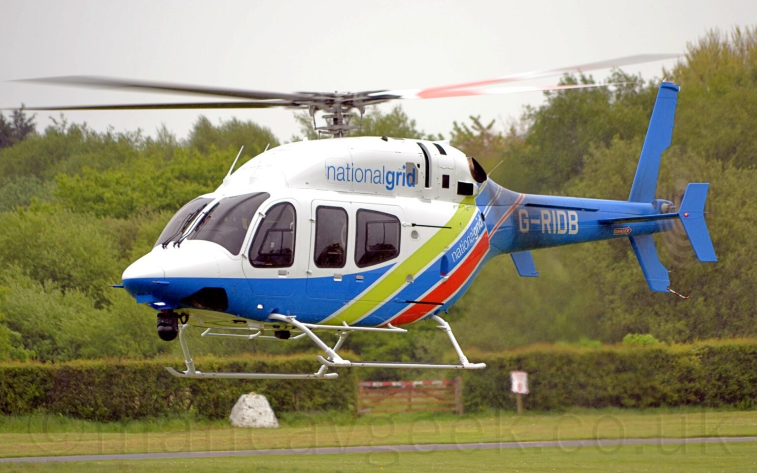 Side view of a helicopter flying from right to left at a very low altitude, about 5 metres above the ground. It is white upper surfaces on the front of the body, with light blue "National Grid" titles on the side of the engine bay on top of the fuselage. The bottom is sky blue, sweeping up towards the rear of the fuselage, and filling the tail boom, with diagonal lime green and red stripes on the rear fuselage, and the white registration "G-RIDB" on the side of the tail boom. Green grass fills the bottom of the frame, with a row of bushes across the middle with a wooden gate with a red and white striped top bar in the centre. Trees fill most of the rest of the frame, with grey sky at the top.