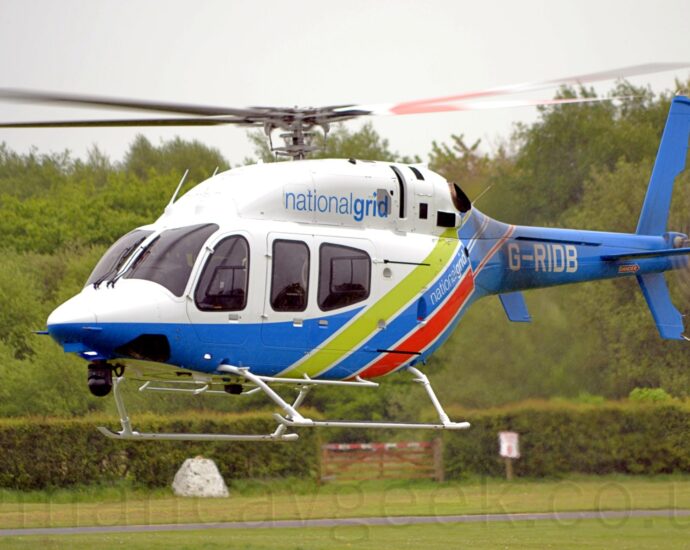 Side view of a helicopter flying from right to left at a very low altitude, about 5 metres above the ground. It is white upper surfaces on the front of the body, with light blue "National Grid" titles on the side of the engine bay on top of the fuselage. The bottom is sky blue, sweeping up towards the rear of the fuselage, and filling the tail boom, with diagonal lime green and red stripes on the rear fuselage, and the white registration "G-RIDB" on the side of the tail boom. Green grass fills the bottom of the frame, with a row of bushes across the middle with a wooden gate with a red and white striped top bar in the centre. Trees fill most of the rest of the frame, with grey sky at the top.