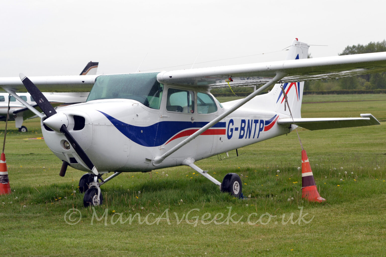 Side front view of a high-winged, single engined light aircraft parked facing to the left in a field of grass. The plane is mostly white, with a blue and red swirly stripe running along the body and up into the tail, overlaid with the blue registration "G-BNTP" on the side of the rear fuselage. An orange traffic cone, filled with weights, is attached to the underside of each wing, weighing it down in case of high winds. Another plane can be seen in the background on the left, under this planes wing, with more grass, and trees in the distance visible under the wing on the right. Flat white sky fills the rest of the frame.