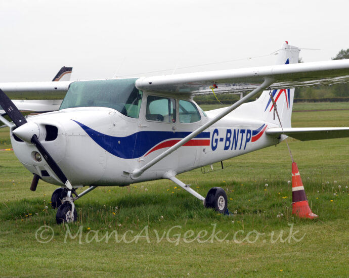 Side front view of a high-winged, single engined light aircraft parked facing to the left in a field of grass. The plane is mostly white, with a blue and red swirly stripe running along the body and up into the tail, overlaid with the blue registration "G-BNTP" on the side of the rear fuselage. An orange traffic cone, filled with weights, is attached to the underside of each wing, weighing it down in case of high winds. Another plane can be seen in the background on the left, under this planes wing, with more grass, and trees in the distance visible under the wing on the right. Flat white sky fills the rest of the frame.