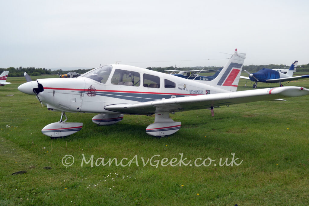 A single-engined light aircraft parked facing to the left in the middle of a grass field. The plane is mostly white, with a pair of thin grey stripes starting from the engine cowling, running backwards and getting wider as it moves rearwards, under a single, thin red stripe, with a similar design on the spats over the undercarriage. The text "Piper Archer II" is on the upper rear fuselage, just behind the cockpit, followed by a block of text in an Arabic script, and a small Palestinian flag with the words "Free Palestine" across the middle in white and black text. Larger grey and red diagonal bands can be seen on the white tail. Green grass fills the foreground, with several light aircraft parked on more grass in the background, leading up to trees meeting grey sky in the distance.