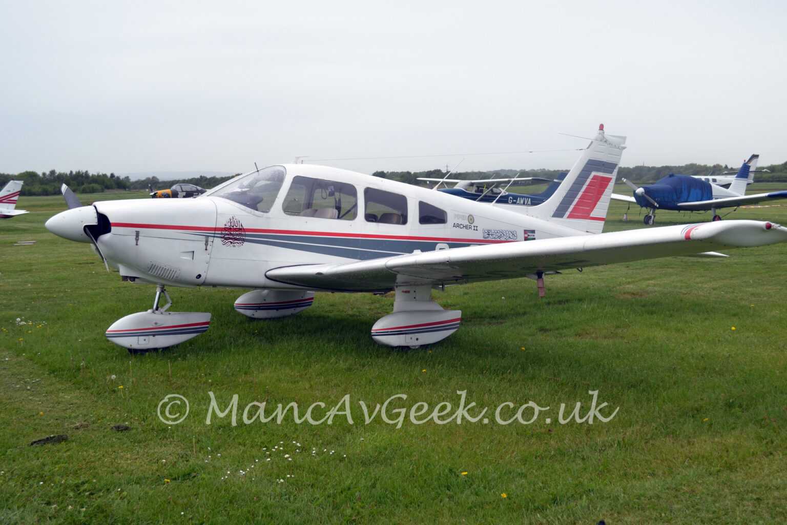 A single-engined light aircraft parked facing to the left in the middle of a grass field. The plane is mostly white, with a pair of thin grey stripes starting from the engine cowling, running backwards and getting wider as it moves rearwards, under a single, thin red stripe, with a similar design on the spats over the undercarriage. The text "Piper Archer II" is on the upper rear fuselage, just behind the cockpit, followed by a block of text in an Arabic script, and a small Palestinian flag with the words "Free Palestine" across the middle in white and black text. Larger grey and red diagonal bands can be seen on the white tail. Green grass fills the foreground, with several light aircraft parked on more grass in the background, leading up to trees meeting grey sky in the distance.