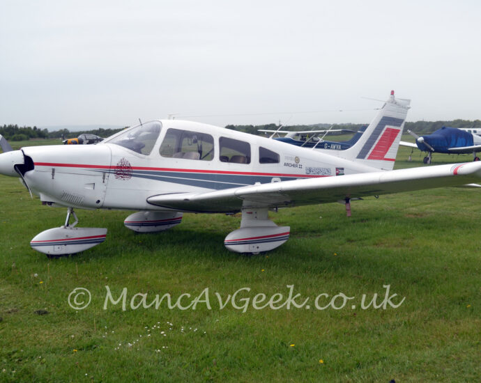 A single-engined light aircraft parked facing to the left in the middle of a grass field. The plane is mostly white, with a pair of thin grey stripes starting from the engine cowling, running backwards and getting wider as it moves rearwards, under a single, thin red stripe, with a similar design on the spats over the undercarriage. The text "Piper Archer II" is on the upper rear fuselage, just behind the cockpit, followed by a block of text in an Arabic script, and a small Palestinian flag with the words "Free Palestine" across the middle in white and black text. Larger grey and red diagonal bands can be seen on the white tail. Green grass fills the foreground, with several light aircraft parked on more grass in the background, leading up to trees meeting grey sky in the distance.