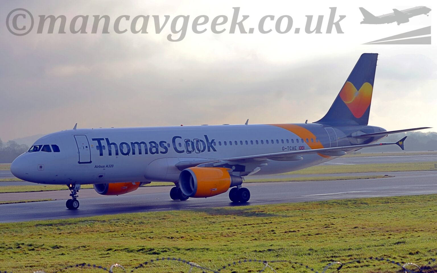 Side view of a twin engined jet airliner taxiing from right to left along a grey taxiway lined with green gr4ass. The plane is mostly white, large, dark grey "Thomas Cook" titles on the upper forward fuselage, partially covering the passenger cabin windows, , with small, grey "Airbus A320" text on the lower forward fuselage, and the grey registration "G-TCAE" next to a small UK flag on the lower rear. A diagonal yellow band wraps around the rear fuselage, next to a light grey band, which is next to a dark grey band which wraps around the tail and the end of the rear fuselage. A large yellow love-heart fills the middle of the tail, with bright yellow matching engine pods mounted under the wings. Grass lines both sides of the taxiway, as well as another taxiway and a runway in the background. Grey sky fills most of the rest of the frame, with a bright spot on the upper middle.