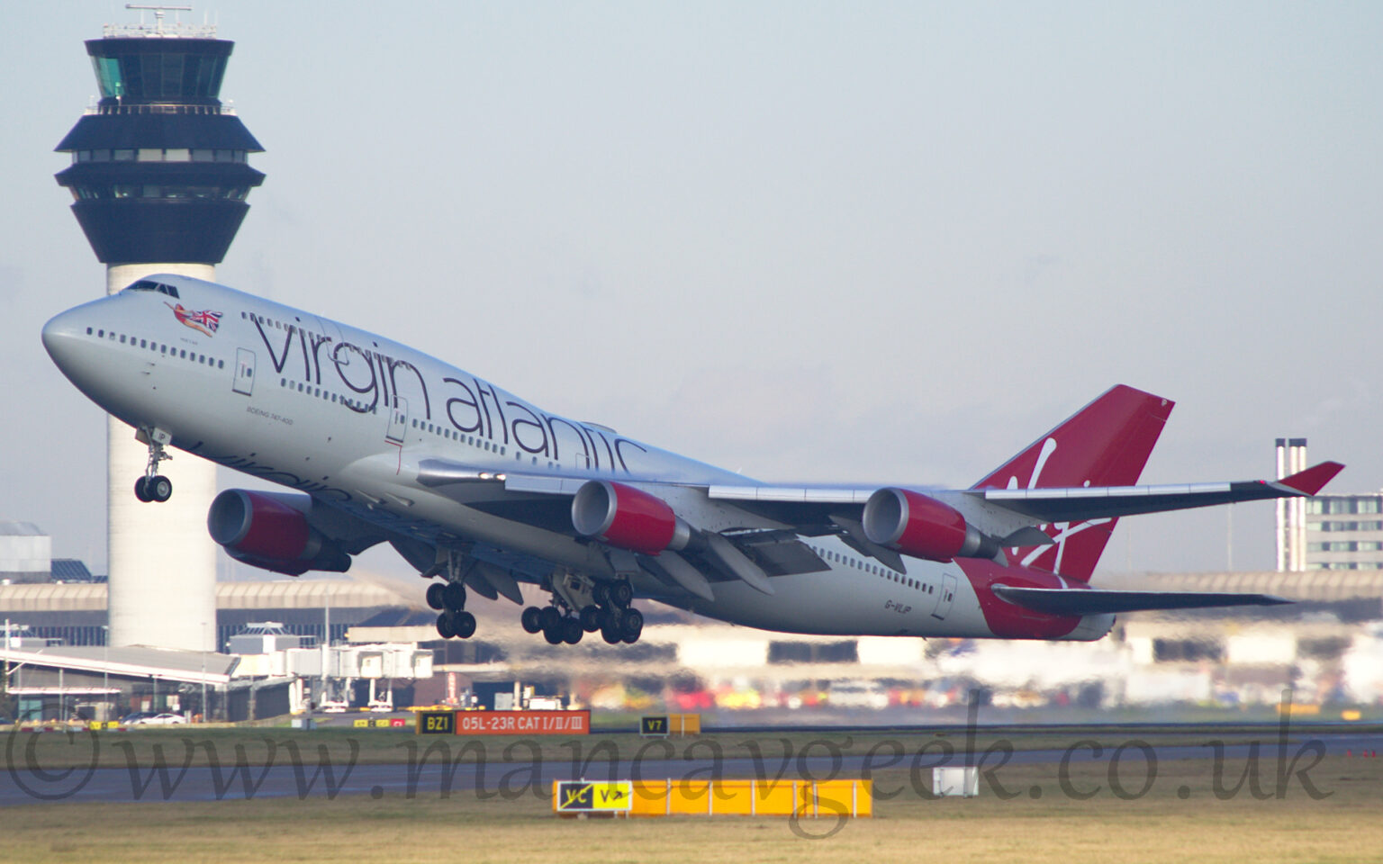 Side view of a very large, 4 engined jet airliner flying from right to left at a very low altitude, with undercarriage extended, flaps deployed from the front and rear of the wings, and the nose raised sharply, suggesting it has just taken off. The plane is mostly white, with large purple "Virgin Atlantic" titles on the upper forward fuselage, and an image of a flying woman wearing a red leotard and a cape made out of a British flag under the cockpit windows. The tail and rear fuselage is a vibrant red, with white, hand-written "Virgin" titles. The engine pods mounted under the wings are the same red, as are the planes up-turned wing-tips. Green grass fills the foreground, while a grey terminal building stretches across the background, with a tall grey tower with black top floors soars into the grey sky on the left.