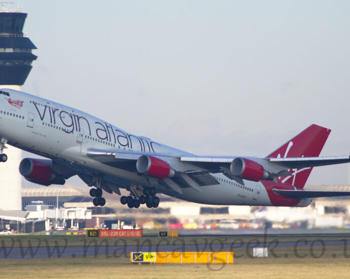 Side view of a very large, 4 engined jet airliner flying from right to left at a very low altitude, with undercarriage extended, flaps deployed from the front and rear of the wings, and the nose raised sharply, suggesting it has just taken off. The plane is mostly white, with large purple "Virgin Atlantic" titles on the upper forward fuselage, and an image of a flying woman wearing a red leotard and a cape made out of a British flag under the cockpit windows. The tail and rear fuselage is a vibrant red, with white, hand-written "Virgin" titles. The engine pods mounted under the wings are the same red, as are the planes up-turned wing-tips. Green grass fills the foreground, while a grey terminal building stretches across the background, with a tall grey tower with black top floors soars into the grey sky on the left.