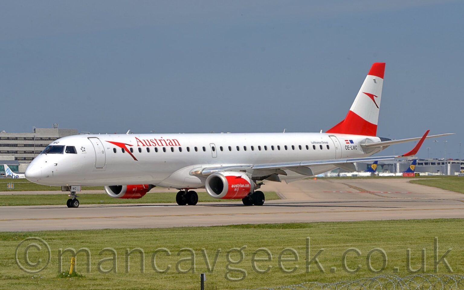 Side view of a white, twin engined jet airliner taxiing from left to right along a grey taxiway. The plane has red "Austrian" titles on the upper forward fuselage, behind a large, red, stylised arrowhead, or possibly a bird. The black registration "OE-LWC" is on the rear fuselage, just aft of the rear door, under small Austrian and EU flags, with grey "Lufthansa Group" titles just ahead of the door. The engine pods under the wings are mostly red, with a white top, and white "AustrianDotCom" text, with the up-turned wing-tips being the same red. The tail is red, with a wide, white, oval band running through the middle, containing a larger version of the arrowhead/bird logo. Green grass fills the foreground, with more grass in the background on the far side of the runway, leading up to grey terminal buildings with the tails of various jet airliners parked outside, under blue sky with hints of grey cloud.