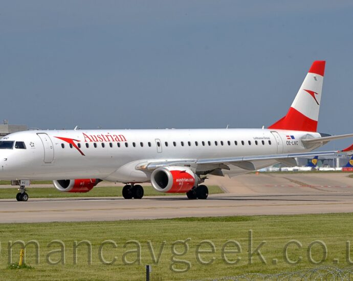 Side view of a white, twin engined jet airliner taxiing from left to right along a grey taxiway. The plane has red "Austrian" titles on the upper forward fuselage, behind a large, red, stylised arrowhead, or possibly a bird. The black registration "OE-LWC" is on the rear fuselage, just aft of the rear door, under small Austrian and EU flags, with grey "Lufthansa Group" titles just ahead of the door. The engine pods under the wings are mostly red, with a white top, and white "AustrianDotCom" text, with the up-turned wing-tips being the same red. The tail is red, with a wide, white, oval band running through the middle, containing a larger version of the arrowhead/bird logo. Green grass fills the foreground, with more grass in the background on the far side of the runway, leading up to grey terminal buildings with the tails of various jet airliners parked outside, under blue sky with hints of grey cloud.