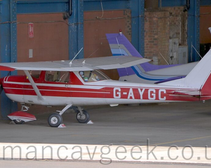 Side view of a single-engined, high-winged light aircraft parked in a brick hangar, facing to the left. The plane is mostly white, with 3 thick red stripes running along the body, and the white registration "G-AYGC" on the side of the rear fuselage. There is an aerodynamic cover on the nose-wheel, with a red top half and white bottom .The tips of the wings and horizontal stabiliser are also painted red. The plane faces a wall made of blue steel support beams, filled in with brick, with some brick sections also covered in brown plaster. The white, light blue, and lilac rear fuselage and tail of another light aircraft can be seen parked facing to the right behind this plane, with the white and blue tail of another beyond that.