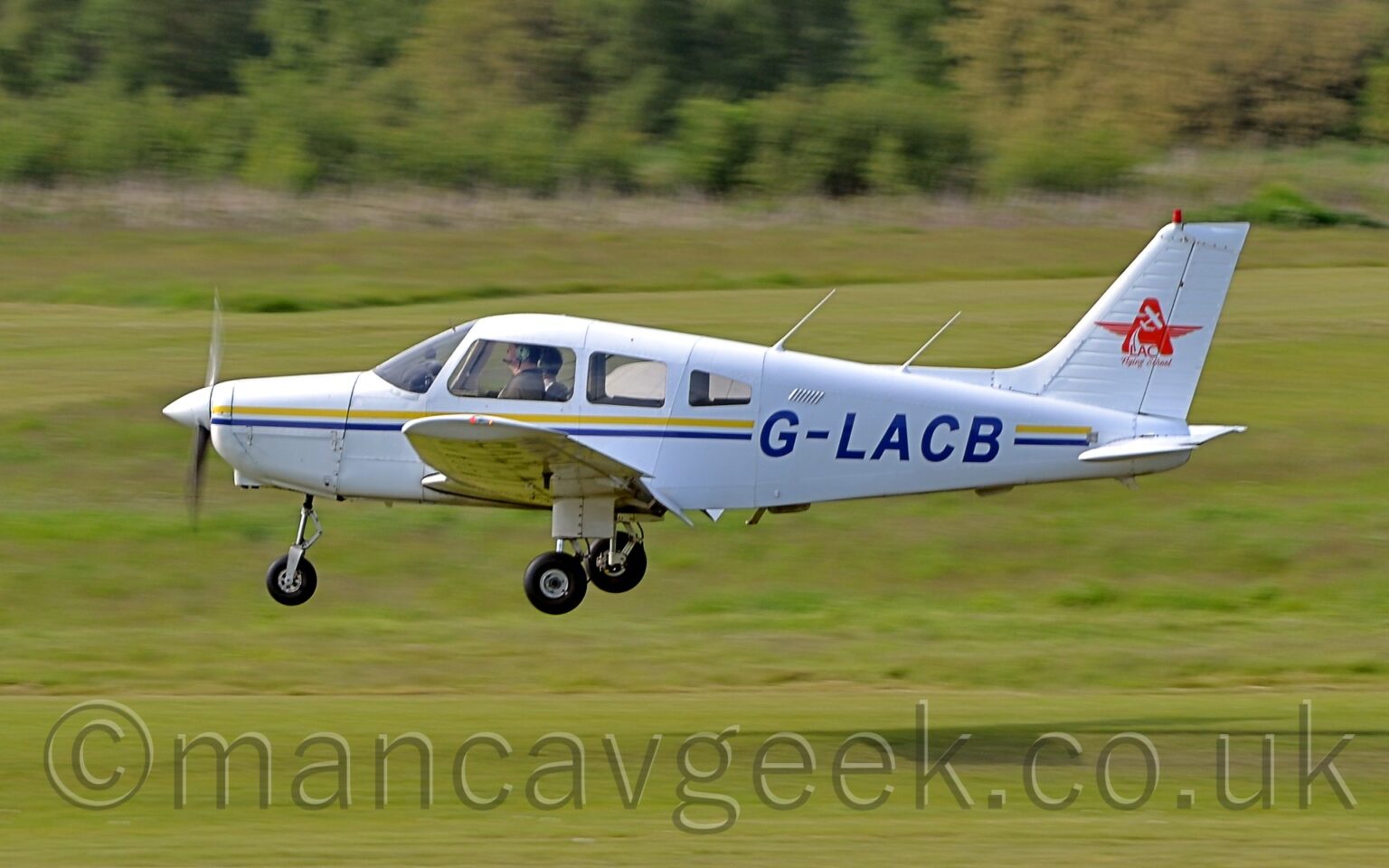 Side view of a single engined light aircraft flying from right to left at a very low altitude, maybe 2 or 3 metres above a grass runway, with flaps extended from the rear of the wings, and flying straight and level, suggesting it is about to land, the fixed undercarriage not telling us anything. The plane is white, with a thin yellow and blue stripe running along the body, overlaid with the blue registration "G-LACB" on the side of the rear fuselage. There is a red star, with the side arms made from a pair of pilots wings, overlaid with an image of a light aircraft soaring into the sky, in the middle of the white tail. Grass fills the foreground and most of the background, with trees and bushes marking the airfield perimeter in the distance.