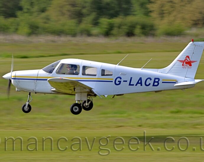 Side view of a single engined light aircraft flying from right to left at a very low altitude, maybe 2 or 3 metres above a grass runway, with flaps extended from the rear of the wings, and flying straight and level, suggesting it is about to land, the fixed undercarriage not telling us anything. The plane is white, with a thin yellow and blue stripe running along the body, overlaid with the blue registration "G-LACB" on the side of the rear fuselage. There is a red star, with the side arms made from a pair of pilots wings, overlaid with an image of a light aircraft soaring into the sky, in the middle of the white tail. Grass fills the foreground and most of the background, with trees and bushes marking the airfield perimeter in the distance.
