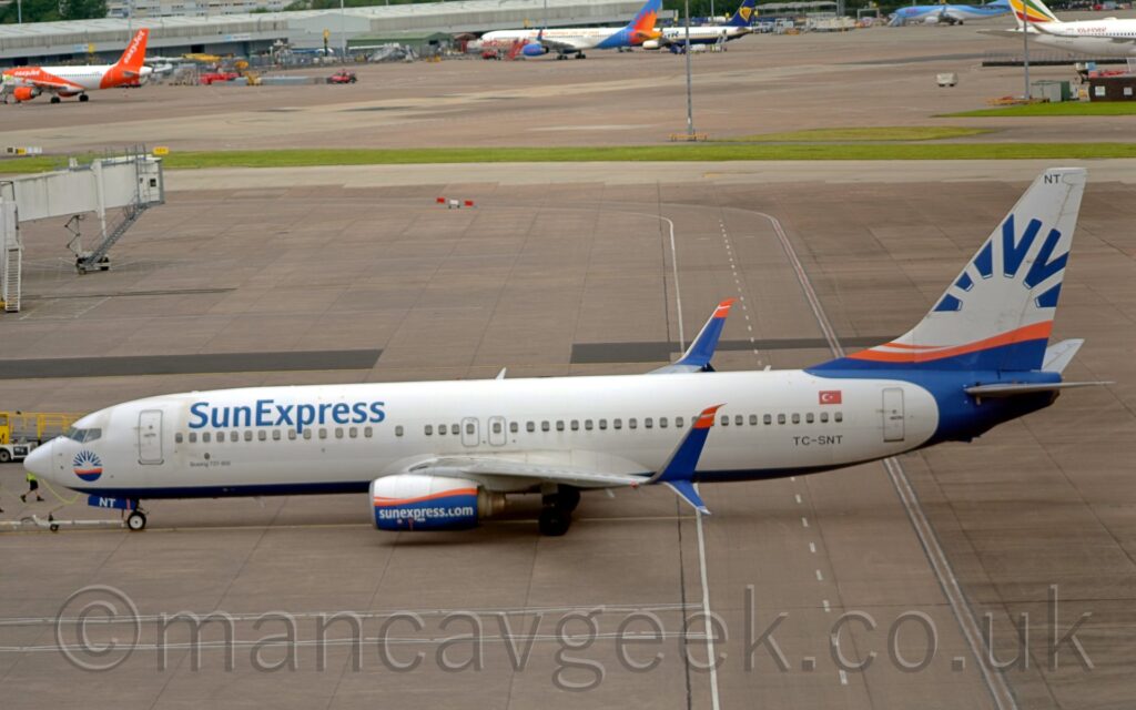 Side view of a twin engined jet airliner facing to the left but being pushed back to the right by an out-of-frame tug attached to the nose-wheel by a long white pole. The plane is largely white, with a dark blue belly, and blue "Sun Express" titles on the upper forward fuselage. The black registration "TC-SNT" is on the lower rear fuselage, below the rearmost passenger cabin windows, with a small Turkish flag just above those windows. A circular, blue sunburst design with an orange band across the middle is on the plane's nose, below the cockpit windows, replicated in a larger form on the tail and rear fuselage. The engine pods under the wings are white on top and blue on the bottom, with an orange stripe across the middle, and white text "SunExpress.com" along the bottom half, while the up-and-down-turned wing-tips are blue, with orange tips on the upper section. Grey concretes apron fills the foreground, with white and yellow markings for aircraft to follow, as well as roadways marked for ground vehicles. More apron fills the background, with a strip of grass separating 2 different areas, with several planes parked against a grey building in the distance.