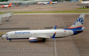 Side view of a twin engined jet airliner facing to the left but being pushed back to the right by an out-of-frame tug attached to the nose-wheel by a long white pole. The plane is largely white, with a dark blue belly, and blue "Sun Express" titles on the upper forward fuselage. The black registration "TC-SNT" is on the lower rear fuselage, below the rearmost passenger cabin windows, with a small Turkish flag just above those windows. A circular, blue sunburst design with an orange band across the middle is on the plane's nose, below the cockpit windows, replicated in a larger form on the tail and rear fuselage. The engine pods under the wings are white on top and blue on the bottom, with an orange stripe across the middle, and white text "SunExpress.com" along the bottom half, while the up-and-down-turned wing-tips are blue, with orange tips on the upper section. Grey concretes apron fills the foreground, with white and yellow markings for aircraft to follow, as well as roadways marked for ground vehicles. More apron fills the background, with a strip of grass separating 2 different areas, with several planes parked against a grey building in the distance.