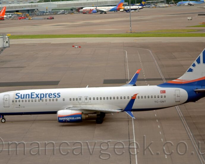 Side view of a twin engined jet airliner facing to the left but being pushed back to the right by an out-of-frame tug attached to the nose-wheel by a long white pole. The plane is largely white, with a dark blue belly, and blue "Sun Express" titles on the upper forward fuselage. The black registration "TC-SNT" is on the lower rear fuselage, below the rearmost passenger cabin windows, with a small Turkish flag just above those windows. A circular, blue sunburst design with an orange band across the middle is on the plane's nose, below the cockpit windows, replicated in a larger form on the tail and rear fuselage. The engine pods under the wings are white on top and blue on the bottom, with an orange stripe across the middle, and white text "SunExpress.com" along the bottom half, while the up-and-down-turned wing-tips are blue, with orange tips on the upper section. Grey concretes apron fills the foreground, with white and yellow markings for aircraft to follow, as well as roadways marked for ground vehicles. More apron fills the background, with a strip of grass separating 2 different areas, with several planes parked against a grey building in the distance.