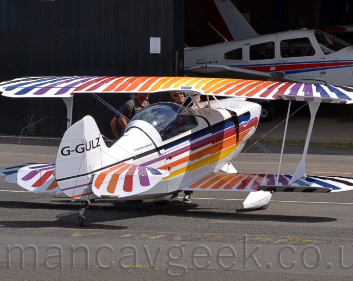 Rear view of a single-engined bi-plane light aircraft parked facing slightly to the right of the camera. The plane is largely white, with black, blue, purple, red, orange, and yellow stripes along the body forming a stylised image of the body of an eagle, with similarly coloured stripes on the top and bottom surfaces of the wings and horizontal stabiliser suggesting wing feathers. There is small black text "Christen Eagle" on the upper rear fuselage, just aft of the large bubble canopy, and the black registration "G-GULZ" on the tail. Three men can be seen standing on the far side of the plane, between the cockpit and engine, with one of them seeming to show the other two something. Grey asphalt apron fills the foreground, with a large black hangar in the background, it's large door open on the right revealing some of the planes stored inside.