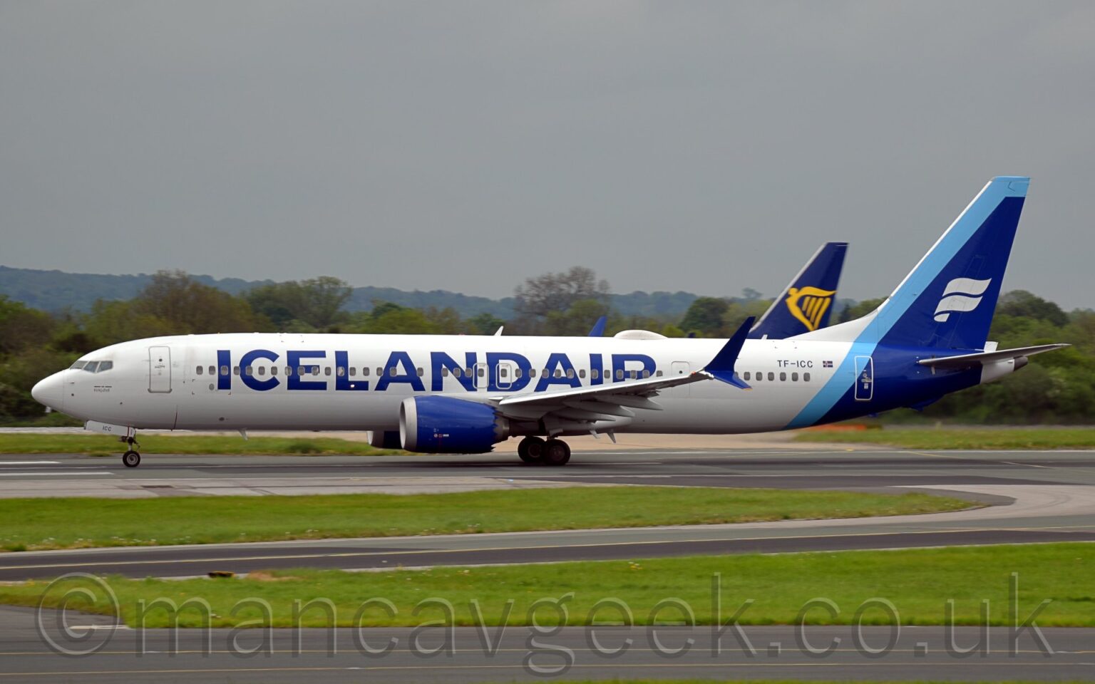 Side view of a twin engined jet airliner moving from right to left at speed along a runway with flaps slightly extended behind the wings, and the nose raised slightly, suggesting it is taking off.The plane is largely white, with large, blue, billboard-style "IcelandAir" titles on the side of the forward fuselage, partially covering the passenger cabin windows. The registration "TF-ICC" is on the upper rear fuselage, next to a small Icelandic flat. A thin light blue stripe wraps around the rear fuselage and the leading edge of the tail, with a thicker dark blue stripe wraps around the tail-cone and the rest of the tail, with a white, waving, double ribbon in the middle. A black taxiway winds it's way through a large grassed area in the foreground, with more grass visible under the planes in the background. The dark blue and yellow tail of another jet airliner is visible over the rear fuselage of this one, with trees beyond that in the distance, under grim grey sky.
