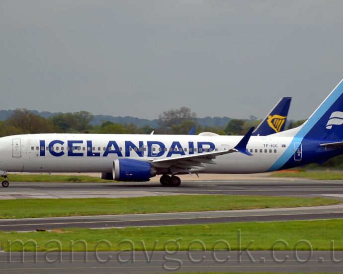 Side view of a twin engined jet airliner moving from right to left at speed along a runway with flaps slightly extended behind the wings, and the nose raised slightly, suggesting it is taking off.The plane is largely white, with large, blue, billboard-style "IcelandAir" titles on the side of the forward fuselage, partially covering the passenger cabin windows. The registration "TF-ICC" is on the upper rear fuselage, next to a small Icelandic flat. A thin light blue stripe wraps around the rear fuselage and the leading edge of the tail, with a thicker dark blue stripe wraps around the tail-cone and the rest of the tail, with a white, waving, double ribbon in the middle. A black taxiway winds it's way through a large grassed area in the foreground, with more grass visible under the planes in the background. The dark blue and yellow tail of another jet airliner is visible over the rear fuselage of this one, with trees beyond that in the distance, under grim grey sky.