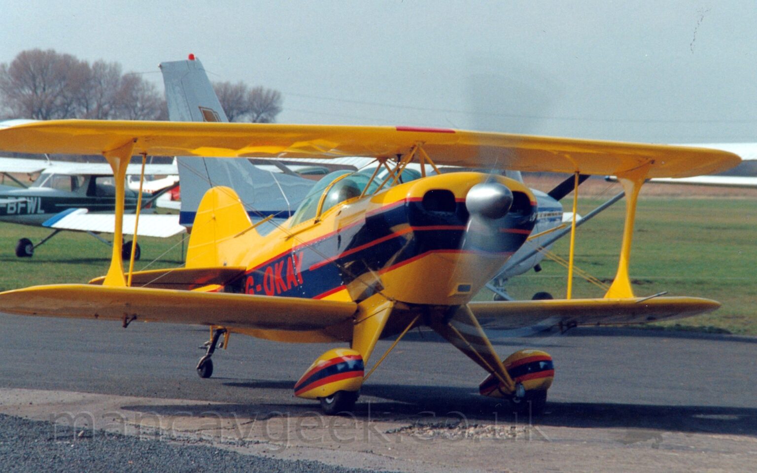Front view of a single-engined bi-plane taxiing from left to right along a black asphalt taxiway, it's propellor in the nose little more than a spinning black blur. The plane is mostly yellow, with a wide black stripe outlined in red , running along the body, overlaid with the red registration "G-OKAY" on the side of the rear fuselage. A thinner black stripe with thicker red outline is on the spats over the main wheels on the front of the body. The pilot can be seen through the large bubble canopy. Green grass fills most of the background, with several light aircraft parked on the left, with trees beyond, under grey-blue sky.