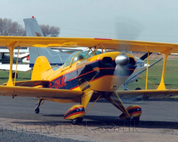Front view of a single-engined bi-plane taxiing from left to right along a black asphalt taxiway, it's propellor in the nose little more than a spinning black blur. The plane is mostly yellow, with a wide black stripe outlined in red , running along the body, overlaid with the red registration "G-OKAY" on the side of the rear fuselage. A thinner black stripe with thicker red outline is on the spats over the main wheels on the front of the body. The pilot can be seen through the large bubble canopy. Green grass fills most of the background, with several light aircraft parked on the left, with trees beyond, under grey-blue sky.