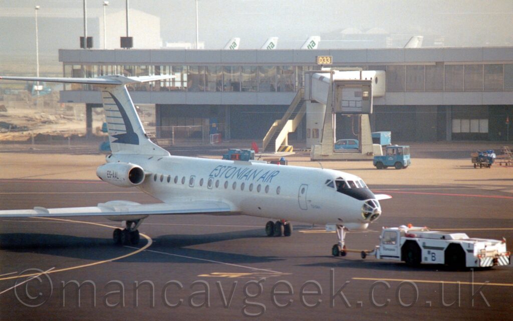 Front side view of a twin engined jet airliner with the engines mounted on the sides of the rear fuselage, facing to the right but being pushed back to the left by a low, white tug attached to the planes nose-wheel by a long white pole. The plane is almost entirely white, with blue "Estonian Air" titles on the upper forward fuselage, and the black registration "ES-AAL" on the side of the white engine pods. The tail is white, with a stylised dark blue flying bird, with thin light blue stripes suggesting speed and movement. The nose cone is heavily glazed, which isn't common on Western airliners. Black apron marked with yellow lines fills the foreground, with more grey apron leading up to a grey terminal building in the background, coming in from the left, with the white tails of more planes visible over the top, under grey sky.