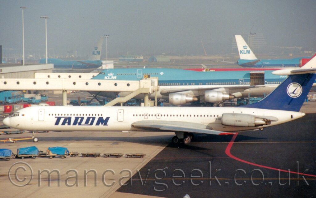 Side view of a white, 4 engined jet airliner, with the grey engines mounted in horizontal pairs on the sides of the rear fuselage, facing to the left but being pushed back to the right by an out-of-frame tug attached to the nose-wheel. There is large, blue text "Tarom" on the sides of the lower forward fuselage, the black registration "YR-IRE" on the side of the rear fuselage just ahead of the engines, and black text "IL-62M" on the side of the engine pods. The tail is blue, with a white circle with a stylised blue bird in the middle. Black taxiway marked with a red line, and grey apron with parked baggage carts, fills the foreground, with a very large, blue and white jet airliner in the background, with a long, thin, white airbridge attached to a terminal building on the left connected to one of the passenger cabin doors, with more similar airliners parked on the left and right of the distance, with everything beyond them being swallowed up in mist.