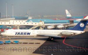 Side view of a white, 4 engined jet airliner, with the grey engines mounted in horizontal pairs on the sides of the rear fuselage, facing to the left but being pushed back to the right by an out-of-frame tug attached to the nose-wheel. There is large, blue text "Tarom" on the sides of the lower forward fuselage, the black registration "YR-IRE" on the side of the rear fuselage just ahead of the engines, and black text "IL-62M" on the side of the engine pods. The tail is blue, with a white circle with a stylised blue bird in the middle. Black taxiway marked with a red line, and grey apron with parked baggage carts, fills the foreground, with a very large, blue and white jet airliner in the background, with a long, thin, white airbridge attached to a terminal building on the left connected to one of the passenger cabin doors, with more similar airliners parked on the left and right of the distance, with everything beyond them being swallowed up in mist.