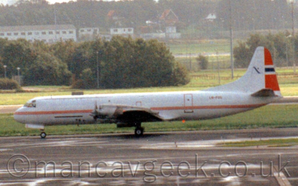 Somewhat grainy side view of a white, 4 propellor-engined cargo plane taxiing from right to left along a grey taxiway.# The plane is almost entirely white, with 3 thin red stripes running along the body, and the red registration "LN-FOG" on the upper rear fuselage. The front of the tail is white, with a white flag with a black diagonal band and a black dot in the top left corner, in the middle. The rear of the tail shows a small part of the Norwegian flag, with red at the top and bottom, a white bar through the middle, and a smaller black bar through the middle of that. Grey apron and taxiways fill the foreground, with grass in the background leading up to trees in the distance, with white houses beyond, and more trees beyond those.