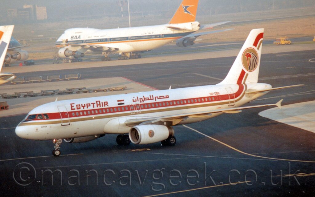 Side view of a white, twin engined jet airliner taxiing from right to left across a black taxiway. The plane has a thick orange stripe running along the body, covering the passenger cabin windows, with a thinner gold stripe below it, both stripes turning up into the tail at the rear. There are orange "EgyptAir" titles on the upper forward fuselage, in English at the front, and in an Arabic script further back, over the wings, with a small Egyptian flag in between. A gold circle is overlaid on the orange and gold stripe on the tail, with the stylised feathered head of a bird facing to the left in the middle, a logo repeated in a smaller form on the sides of the white engine pods under the wings. The black taxiway in the foreground continues into the background, wending it's way around large grey patches of concrete apron, with a very large white and orange , 4 engined jet airliner in the distance, with everything beyond that slowly vanishing in to haze.