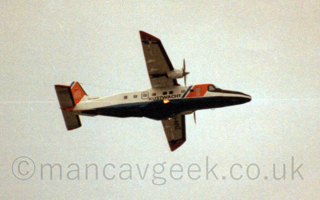 Low side view of a high-winged, twin propellor-engined airliner with a long nose, flying from left to right at a low altitude, with flaps extended slightly from the rear of the wings, and undercarriage retracted, suggesting it has just taken off. The plane is mostly white, withs a thick diagonal orange band followed by a thinner, diagonal blue band, on the forward fuselage, with black "Kustwacht" titles on the lower middle fuselage, and a dark blue belly. Another diagonal orange and dark blue stripe fills most of the tail. The leading edges of the wings and tail are black, and there is a black anti-dazzle panel on the top of the plane's nose, in front of the cockpit. Hazy grey sky fills the rest of the frame.