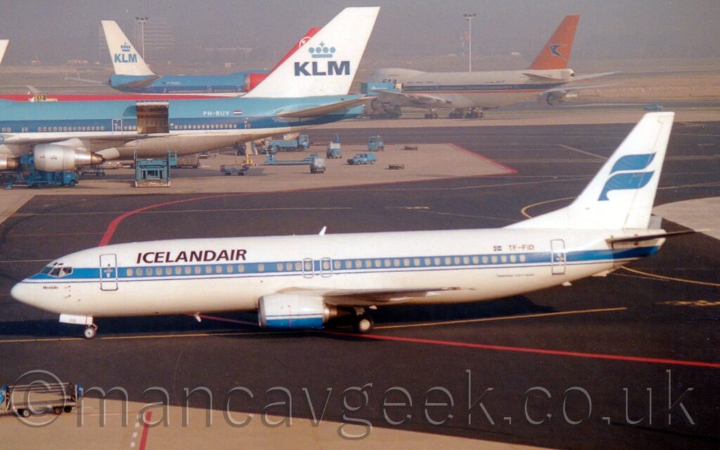 Side view of a white, twin engined jet airliner, following a yellow line on a black taxiway as it taxis from right to left. There are black "IcelandAir" titles on the upper forward fuselage, and a thick, blue stripe running along the body, covering the passenger cabin windows. The black registration "TF-FID" is on the upper rear fuselage, next to a small Icelandic flag, with black text "Boeing 737-400" on the lower rear. The engine pods under the wings are white, with a blue belly. The tail is white, with a stylised blue bird with outstretched wings in the middle. Black taxiway winds it's way through grey concrete apron in the background, with larger jet airliners, some blue and white, one white and orange, one mostly red, in the distance, with buildings being swallowed up in mist right at the back of the frame.