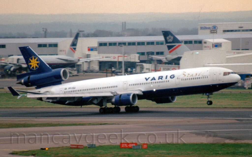 Side view of a 3 engined jet airliner landing heavily on a black runway, with flaps extended behind and in front of the wings the wings, and the rear section of the engine pods slid back, exposing the thrust reversers. The plane is mostly white, with dark blue "Varig" and gold "Brazil" titles on the upper forward fuselage, and the black registration "PP-VQJ" next to a small Brazilian flag on the upper rear. The belly is dark blue, as are the engine pods under the wings, and the elongated centre engine pod running through the base of the blue tail. The tail itself has a large, yellow, 8-pointed compass rose in the middle. A taxiway crosses the runway immediately in front of the plane, both lined with green grass. A large grey terminal building stretches across the background, with a couple of grey and dark blue jet airliners parked against this terminal. In the distance, trees and buildings slowly vanish into bright but hazy skies.