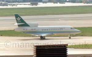 Side view of a 3 engined BizJet taxiing from left to right along a grey taxiway. The plane is largely white, with a green and blue stripe running along the body, and black "Saudi Arabian Airlines" titles, in both English and Arabic, on the upper forward fuselage. The black registration "HZ-AFT" is on the side of the white engine pods mounted on the sides of the upper rear fuselage, just below the centre engine air intake. The green tail has a white logo, an inverted triangle with green crossed sabre, under a white palm tree, is in the middle, over white "Saudia" text. A ventilation stack is in the middle of the roof of a building at the bottom of the frame, with grey apron behind that, filling the foreground. A wide, black runway stretches across the background, with grass on the far sides, with more apron filled with cargo containers in the distance.