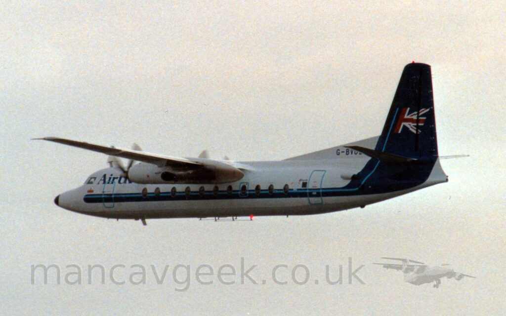 Side view of a high-winged, twin propellor-engined airliner slying from right to left at a low altitude, with flaps slightly extended from the rear of the wings and undercarriage stowed away, and the nose raised, suggesting it has just taken off. The plane is largely white, with a dark blue stripe running along the body, sweeping up to fill the tail, with a thinner, pale blue stripe running through that, acting as a pole against which half a UK flag flutters on the tail. Flat grey sky fills the rest of the frame.
